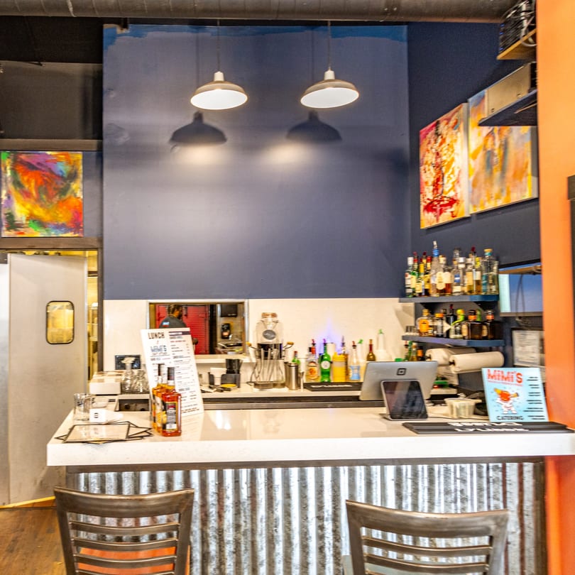 MiMi's Craft Kitchen bar area with bottles, menus, and abstract art on a dark blue wall.