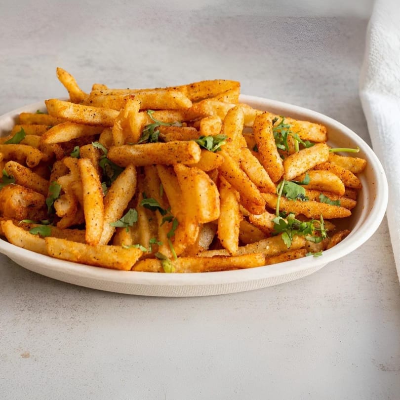 A white bowl filled with golden-brown seasoned fries and fresh green herbs, on a light grey surface.