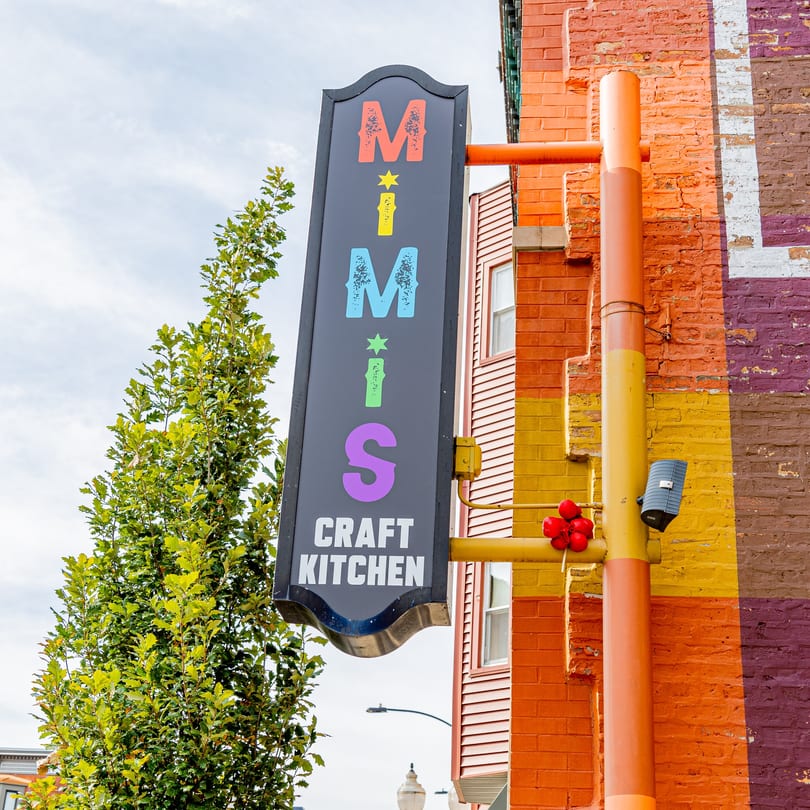 MiMi's Craft Kitchen sign, with colorful letters, attached to a brick building with a painted mural.