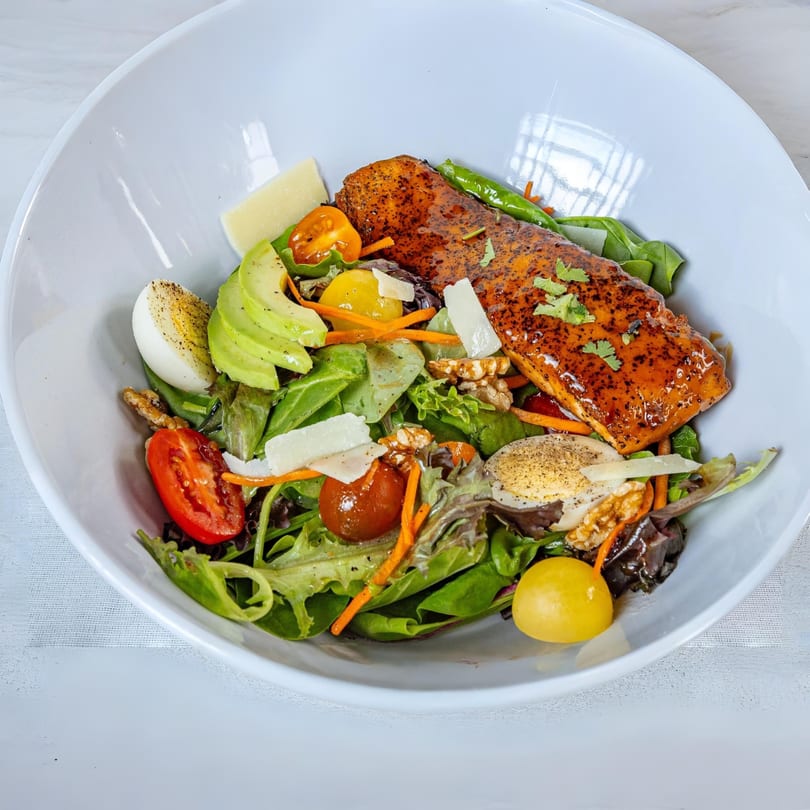 A MiMi's Craft Kitchen salmon salad with avocado, eggs, tomatoes, and greens in a white bowl.
