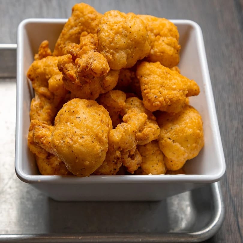 A white square bowl filled with golden-brown, crispy fried cauliflower bites on a metal tray.